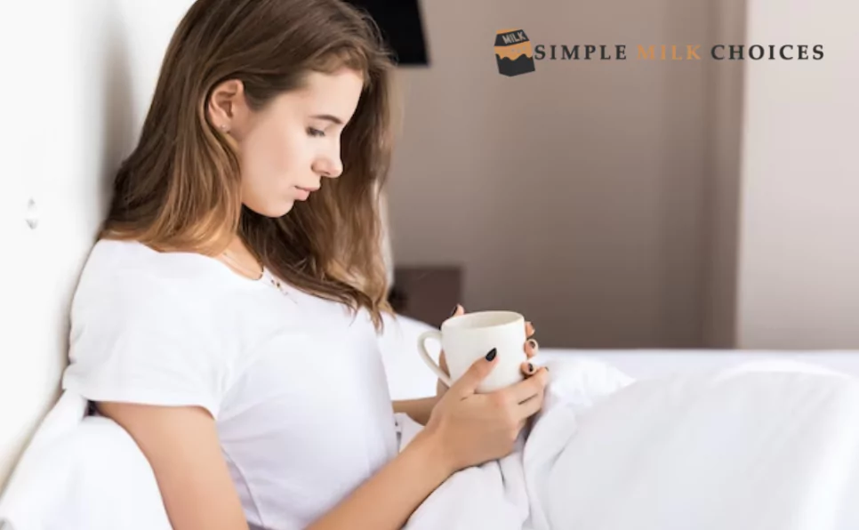 An attentive young girl sits cross-legged on a neatly made bed, her gaze fixed on a glass of creamy almond milk resting on a coaster. The soft glow of the bedside lamp illuminates the scene, highlighting the tranquil moment captured in the quiet bedroom.