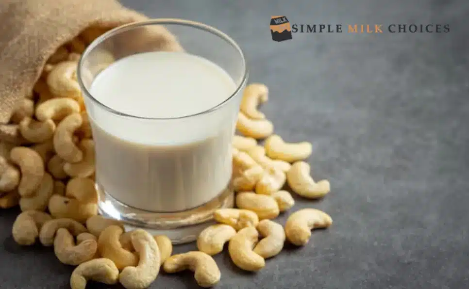 Glass of creamy cashew milk on a wooden surface with scattered cashew nuts around.